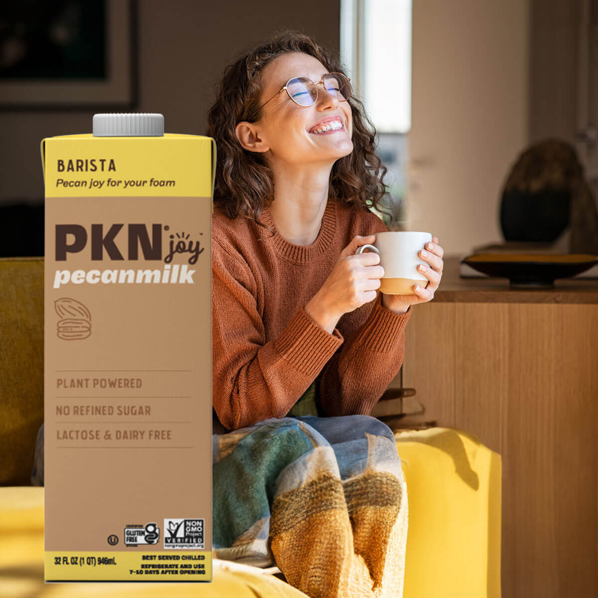 Smiling woman enjoying a hot drink on a couch with PKN Joy Barista pecan milk carton, highlighting plant-based, dairy-free milk for creamy foam.