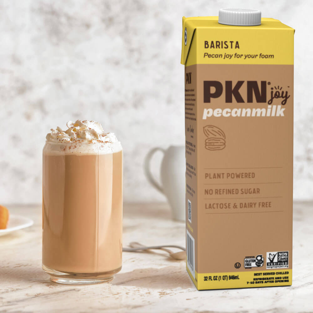 PKN Joy Barista pecan milk carton beside a creamy latte topped with foam, highlighting plant-based, dairy-free milk ideal for barista-style drinks.