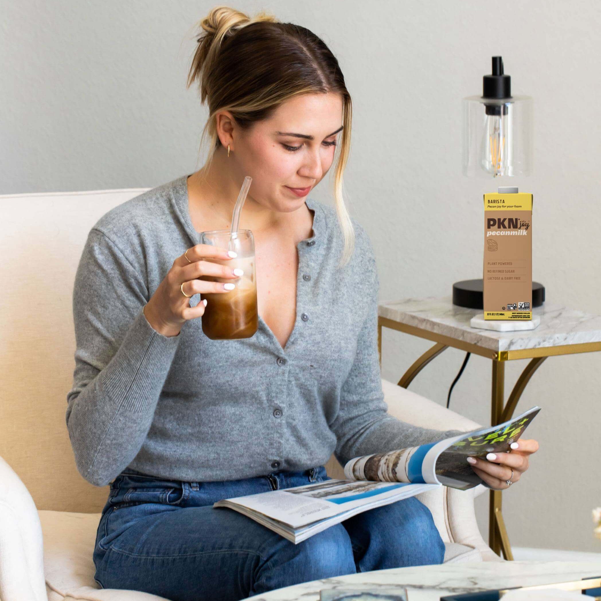 Woman reading a magazine while sipping iced coffee, with PKN Joy Barista pecan milk carton on a side table, highlighting plant-based barista blend.