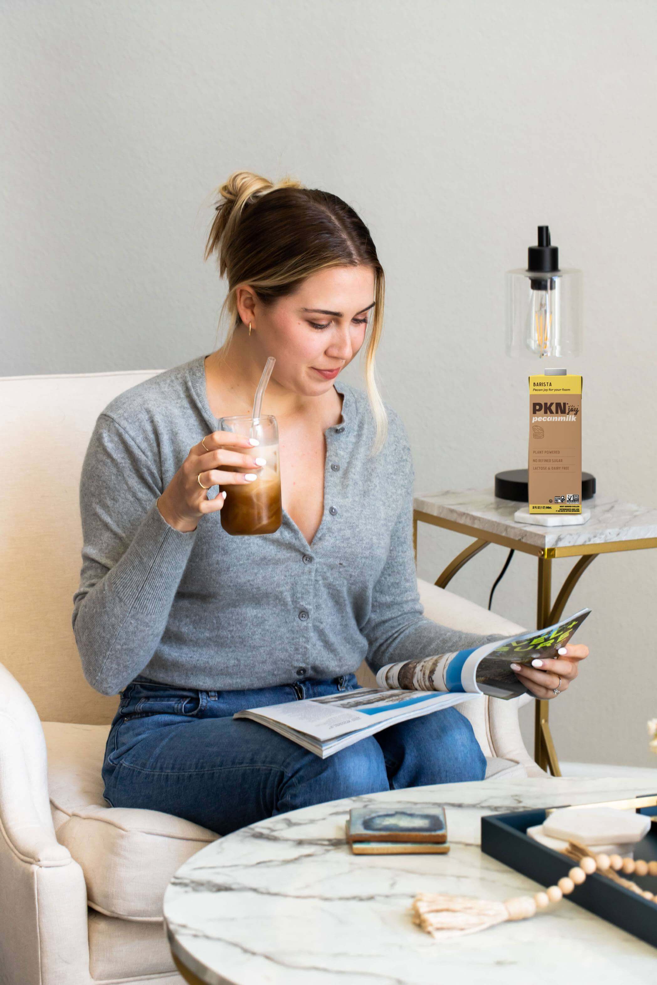 Woman reading a magazine while sipping iced coffee, with PKN Joy Barista pecan milk carton on a side table, highlighting plant-based barista blend.
