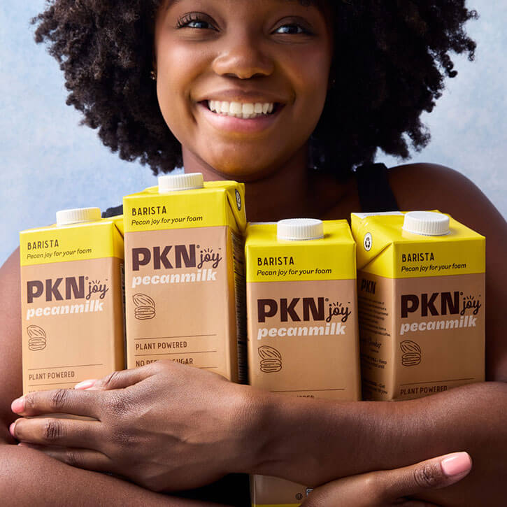 Smiling woman holding multiple PKN Joy Barista pecan milk cartons, highlighting plant-based, dairy-free milk designed for creamy foam.