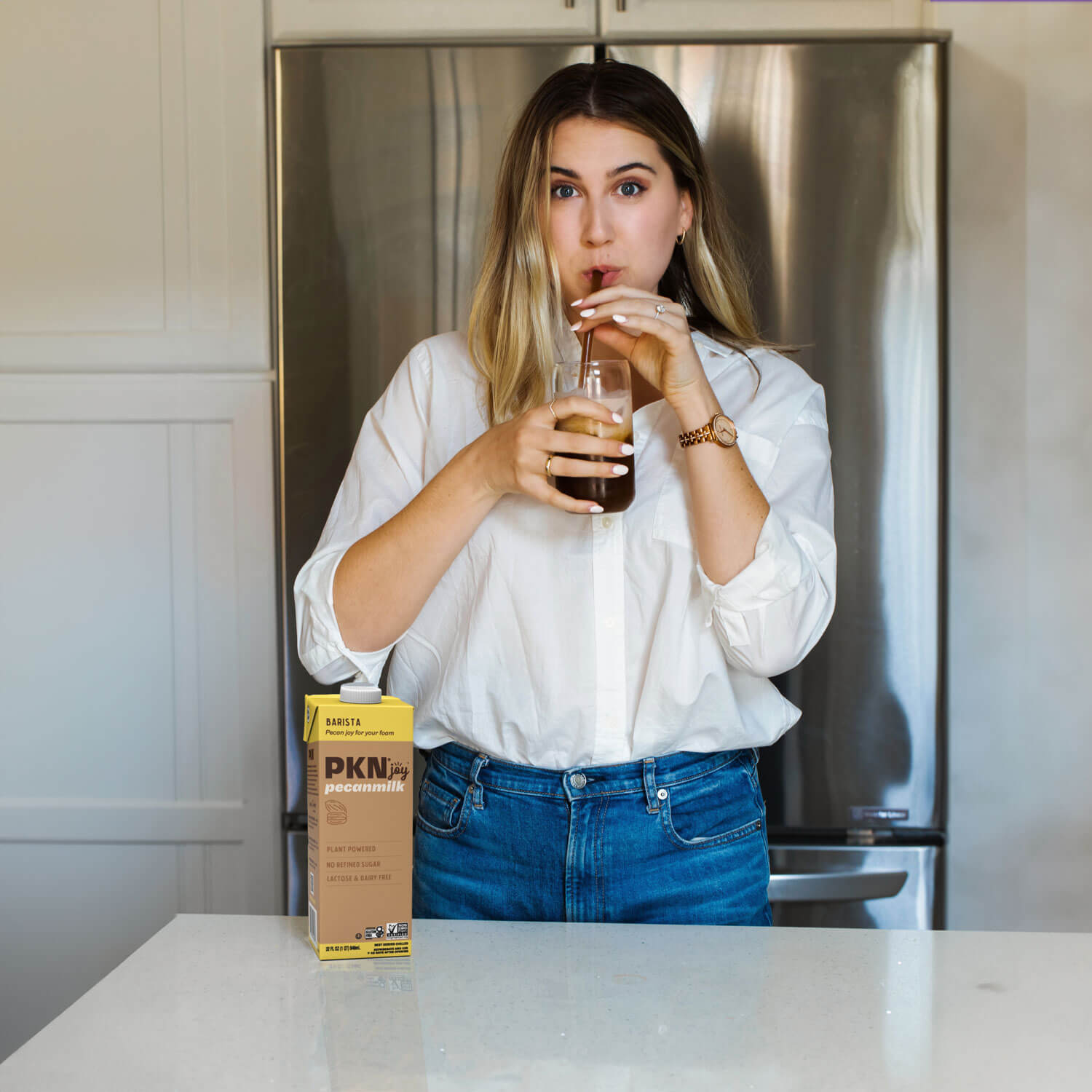 Woman sipping iced coffee with a straw in a kitchen, with PKN Joy Barista pecan milk carton on the counter, highlighting plant-based barista blend.