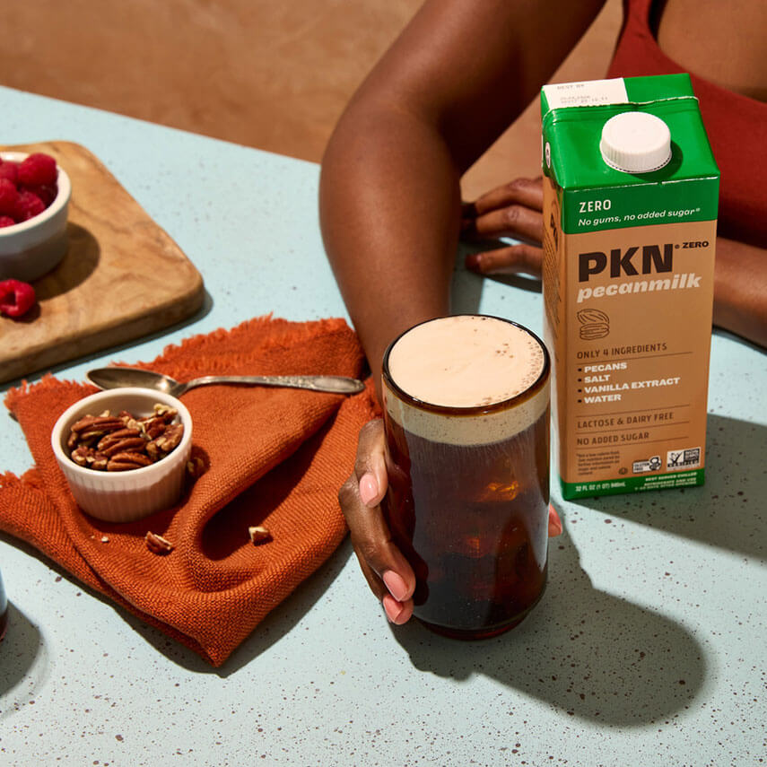 Hand holding iced coffee topped with pecan milk foam beside PKN Zero pecan milk carton, with pecans and raspberries on a colorful kitchen counter.
